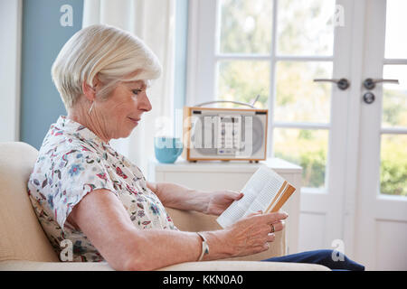 Senior donna seduta in poltrona a leggere un libro a casa Foto Stock