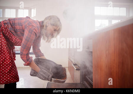 Donna di mezza età apertura piena di fumo forno in cucina Foto Stock