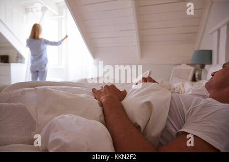 La donna si apre le tende e guarda fuori della finestra come uomo giace nel letto Foto Stock