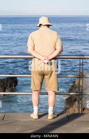 Uomo vestito con cappello, camicia e pantaloncini beige che si affacciano sul mare. Foto Stock