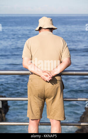 Uomo vestito con cappello, camicia e pantaloncini beige che si affacciano sul mare. Foto Stock