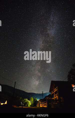 La Via Lattea oltre le montagne e il villaggio su una notte d'estate. Casa in legno di tre piani in primo piano. Foto Stock