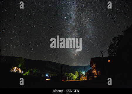 La Via Lattea oltre le montagne e il villaggio su una notte d'estate. Casa in legno di tre piani in primo piano. Foto Stock