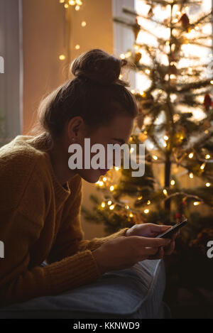 Donna che utilizza il suo telefono cellulare vicino a un albero di Natale a casa Foto Stock