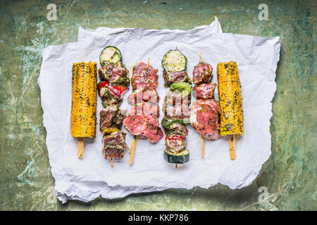 Varie gli spiedini di carne per grill con verdure , le pannocchie e condimento su verde sfondo rustico , vista superiore Foto Stock