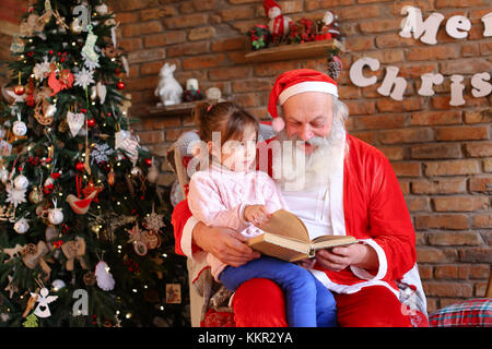 Curioso bambino femmina ascolta con attenzione alla storia interessante da leggere il libro di Babbo Natale in accoglienti e spaziose camere decorate per nuovi anni holi Foto Stock