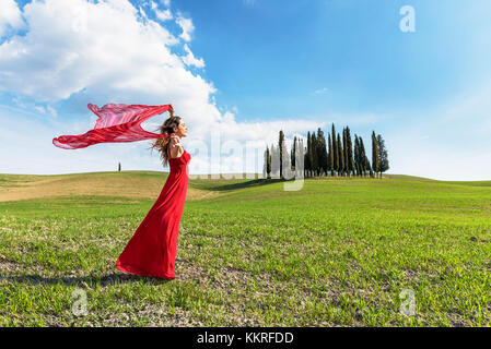 San Quirico d'Orcia, Val d'Orcia, Siena, Toscana, Italia. Una giovane donna in abito rosso rilassante in un campo di grano vicino i cipressi della Val d' Orcia Foto Stock