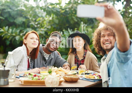 Gruppo di amici internazionali guardando la fotocamera dello smartphone mentre si effettua selfie servita dalla tabella Foto Stock