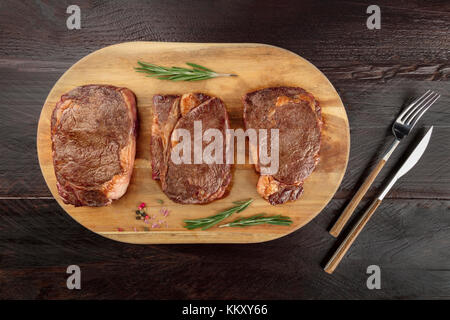 Una foto aerea di tre fette di carne cotta, filetti di manzo, ripresa dall'alto sul buio rustica con rametti di rosmarino, sale, pepe e un p Foto Stock