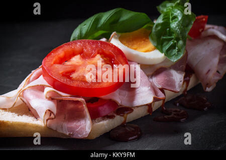 Sandwich con prosciutto, uova, ortaggi e ketchup su sfondo nero. Vedere serie Foto Stock