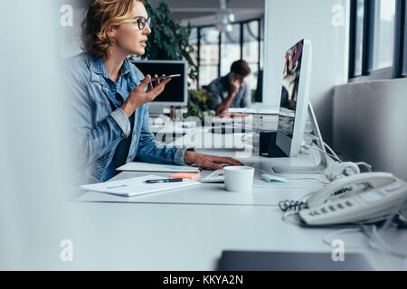 Giovane imprenditrice parlando al telefono con altoparlante e di lavoro sul computer. Giovane donna seduta davanti al monitor con smart phone. Foto Stock