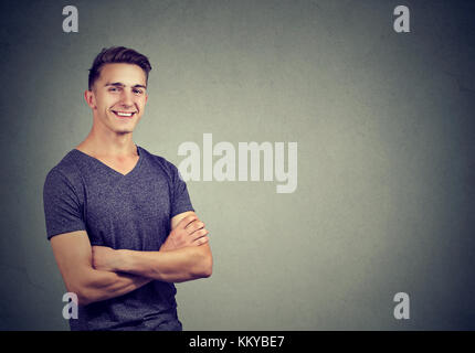 Casualmente bello fiducioso giovane uomo in t-shirt mantenendo le braccia incrociate mentre in piedi contro la parete sullo sfondo Foto Stock