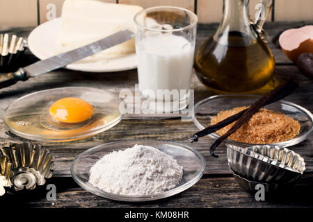 Ingredienti per la cottura (farina, l'uovo, lo zucchero di canna, latte) con l'annata tortina di moduli sul vecchio tavolo in legno Foto Stock