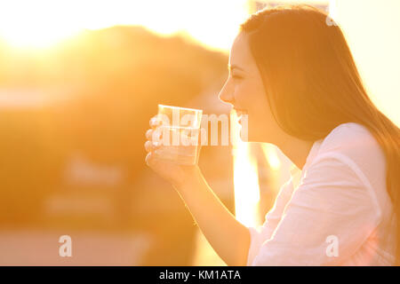 Vista laterale il ritratto di una donna felice con in mano un bicchiere di acqua al di fuori di un balcone al tramonto Foto Stock