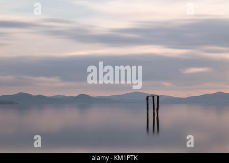 Una visione minimalista di un lago al tramonto, con luce tenue e di toni e pali in primo piano riflettendo sull'acqua Foto Stock