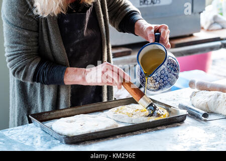 Una femmina di baker si prepara la focaccia italiana pane piatto in una stalla all'aperto ella è versando olio sulla parte superiore dell'impasto prima di essa è collocata in un forno per cucinare Foto Stock