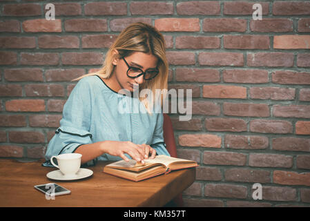 Giovane donna bionda seduta godetevi la lettura di prenotare presso il cafe. vintage nei toni del filtro. Foto Stock