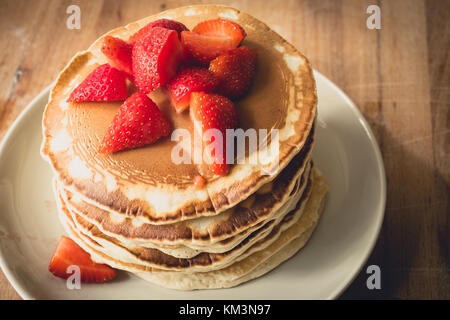 Una pila di frittelle fatte in casa su una piastra bianca con fragole tagliate nella parte superiore. Formato orizzontale. Foto Stock