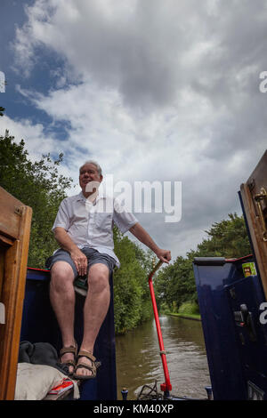 Un uomo in pantaloncini e maglietta a maniche corte si trova nella parte posteriore di un narrowboat mentre lo sterzo con il timone, Coventry canal, Staffordshire, Inghilterra Foto Stock