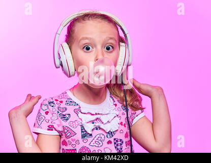 Bambina con le cuffie che soffia su una bolla di gomma su un sfondo rosa Foto Stock