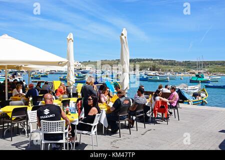 I turisti in un momento di relax a un cafe' sul marciapiede lungo il lungomare con le tradizionali barche da pesca maltesi nel porto verso la parte posteriore, Marsaxlokk, malta, europ Foto Stock