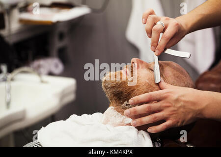 Un cliente seduto al barbiere di sedia, avente una rasatura bagnata da un barbiere utilizzando una gola tagliata rasoio. Foto Stock