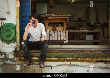 Un uomo in un teeshirt seduto su una fase di bere da un bicchiere, tenendo il suo smart phone. Foto Stock