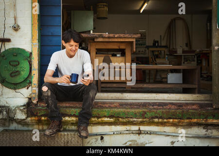 Un uomo in un teeshirt avente un tea break, seduto sui gradini di un laboratorio di controllo il suo telefono. Foto Stock