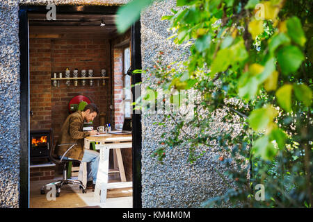 Vista attraverso una porta aperta, un designer seduto nel suo marocchineria workshop ad una scrivania, disegno woodburning stufa con un incandescente Fire lit. Foto Stock