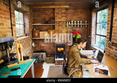 Un designer seduto nel suo marocchineria officina, a una scrivania utilizzando un laptop. woodburning stufa con un incandescente Fire lit. Foto Stock