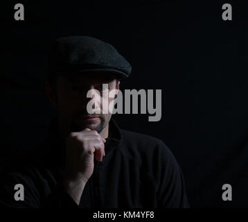 Ritratto artistico dell'uomo isolato con cappuccio scuro, viso mezzo-illuminato, con la telecamera, mano sul mento, sotto i riflettori. L'illuminazione sottile crea un'immagine ombreggiata. Foto Stock