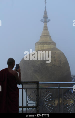 Foto da dietro del monaco prendendo foto sul telefono. il golden rock pagoda si siede sul monte kyaiktiyo precariamente arroccato sulla scogliera egde. ricoperto in oro Foto Stock