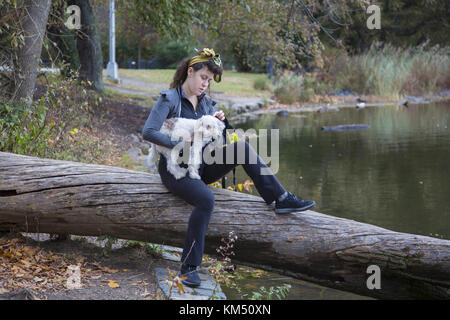 Giovane donna con il suo compagno canino è situato lungo il lago in un giorno di autunno caldo in Prospect Park di Brooklyn, New York. Foto Stock