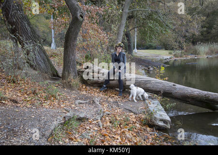 Giovane donna con il suo compagno canino è situato lungo il lago in un giorno di autunno caldo in Prospect Park di Brooklyn, New York. Foto Stock