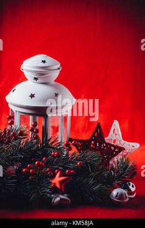 Bianco Natale lanterna è in piedi con una candela che brucia in essa con un abete del ramo e maglia stelle nella parte anteriore di esso su uno sfondo di colore rosso. il natale fa Foto Stock