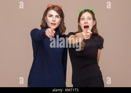 Belle Donne in piedi accanto a ciascun altro e puntamento dita alla fotocamera e shock. studio shot, luce sfondo marrone Foto Stock