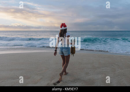 Donna che indossa un cappello di Natale che cammina sulla spiaggia portando un ananas, Haleiwa, Hawaii, Stati Uniti Foto Stock