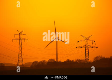 Di elettricità ad alta tensione tralicci / torri di trasmissione e la turbina eolica stagliano tramonto Foto Stock