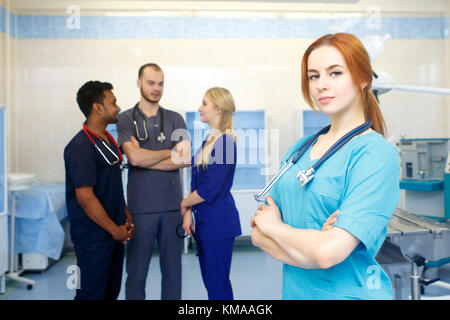 Medico donna nella parte anteriore del team, guardando la telecamera con il team medico in background. multirazziale team di giovani medici in un ospedale in piedi in una sala operatoria. Foto Stock