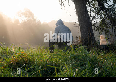 Problemi di rapporto con l immagine di un uomo e un orso di peluche seduto su una panca in legno sotto un albero, mentre godendo la nebbia di mattina e raggi solari. Foto Stock