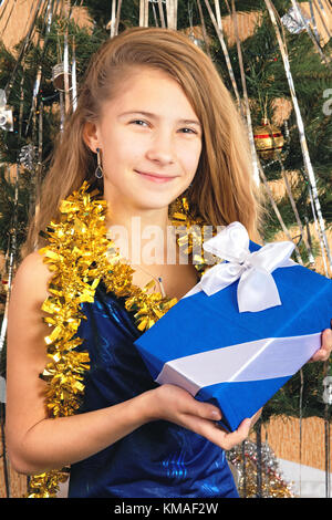 Bella ragazza teen vicino albero di natale tenendo un dono-box di colore blu fasciata con nastro Foto Stock