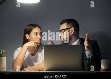 Barbuto corporate manager in tuta discutere le idee con la millenaria lavoratore di sesso femminile che a turno di notte. business datori di lavoro seduto alla scrivania dietro il laptop. Foto Stock