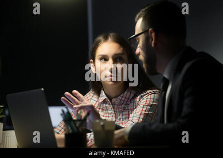 Giovani intern ascolto di guida dando istruzioni seduto alla scrivania a tarda sera, manager femmina insegnamento partecipante con il computer portatile in ufficio interno, col Foto Stock