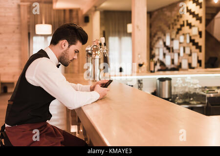 Cameriere seduto al banco bar con lo smartphone Foto Stock