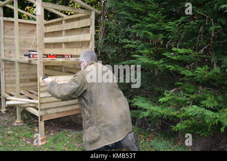 Capelli grigi giardiniere maschio in antichi verde cerato barbour jacket la costruzione di un registro di legname store in giardino da trattati featheredge imbarco e legno Foto Stock