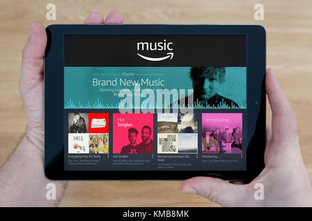 Un uomo guarda al Amazon Sito web musica sul suo iPad dispositivo tablet, sparato contro un tavolo di legno top sfondo (solo uso editoriale) Foto Stock