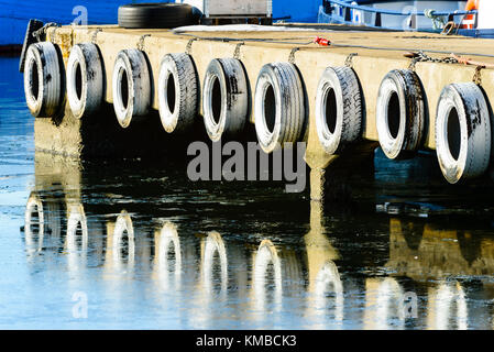 Riga del dipinto di bianco di pneumatici per auto appeso come paraurti o parafanghi sul Quayside. acqua ghiacciata di seguito riflette i pneumatici. Foto Stock