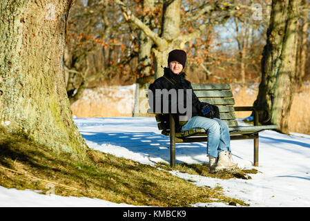 Giovane femmina adulta vestita di nero e blu, in appoggio su una panchina nel parco in inverno o in primavera. neve sulla terra. soleggiata giornata calda con alcuni disgelo aroun Foto Stock