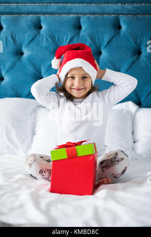 Felice esce da bambina santa's hat azienda confezione regalo, guardando la fotocamera mentre è seduto sul letto Foto Stock