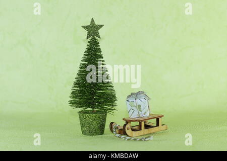 Una miniatura in legno portante della slitta in un piccolo regalo accanto ad un luminoso verde albero di natale su uno sfondo verde chiaro Foto Stock